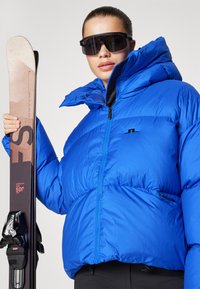 Modra puffer jakna z visokim ovratnikom, svetleča tekstura in črn logotip. Model drži smučko s temnimi vezmi, nosi prevelike črne smučarske očale.