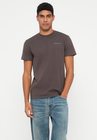 Mężczyzna w ciemno-brązowej koszulce Tommy Jeans i jasnoniebieskich dżinsach, stojący z jedną ręką w kieszeni na tle jasnej białej ściany.