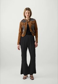 Jonge vrouw staat rechtop, gekleed in een bruine leren cropped jas, zwart top, zwarte high-waisted flared broek en zwarte spitse hakken, tegen een effen achtergrond.