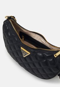 Musta tikattu nahkalaukku, jossa on kullankeltainen kolmionmuotoinen logo, kultainen vetoketju ja vuorattu sisusta erillisellä vetoketjullisella taskulla.