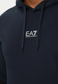 Felpa navy in tessuto morbido, con cappuccio regolabile e logo frontale "EA7 EMPORIO ARMANI" in bianco.