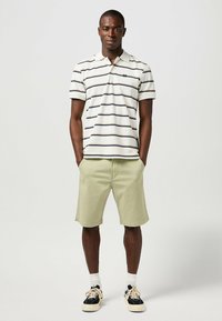 Polo a righe bianche con righe blu navy e rosso scuro, abbinato a pantaloni corti verde chiaro. Sneakers nere con accenti beige e calze bianche alla caviglia.