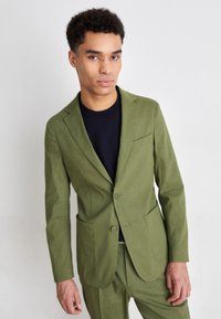 Giacca da abito verde in cotone con chiusura a un bottone, rever a letto, una tasca sul petto e tasche laterali; abbinata a una camicia scura.