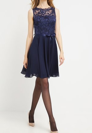 Femme portant une robe sans manches bleu marine avec un corsage en dentelle, un ruban de satin à la taille, une jupe transparente, des collants noirs et des talons hauts beiges, debout sur un fond blanc.