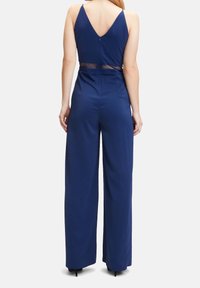 Marinblå jumpsuit med V-rygg, tunna remmar och genomskinlig midjedetalj. Vidbenad design, slät textur och dragkedjestängning på baksidan.