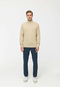 Beige garbós pulóver hosszú ujjakkal, sötétkék nadrággal párosítva. A modellen fehér és szürke sportcipők vannak, egy egyszínű háttér előtt áll.