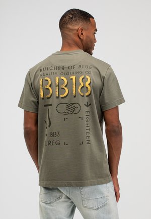 Mann in olivgrünem T-Shirt mit großem gelbem und schwarzem Text und Grafiken auf der Rückseite, trägt hellblaue Jeans, vom Kamerablick abgewandt.