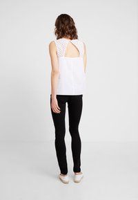 Top blanco con detalles de encaje en los hombros y un corte en la espalda, combinado con pantalones negros. La modelo está de espaldas a la cámara, con los zapatos visibles.