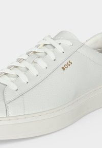 Baskets en cuir blanc avec lacets blancs et logo doré "BOSS" sur le côté, avec une semelle épaisse en caoutchouc blanc.