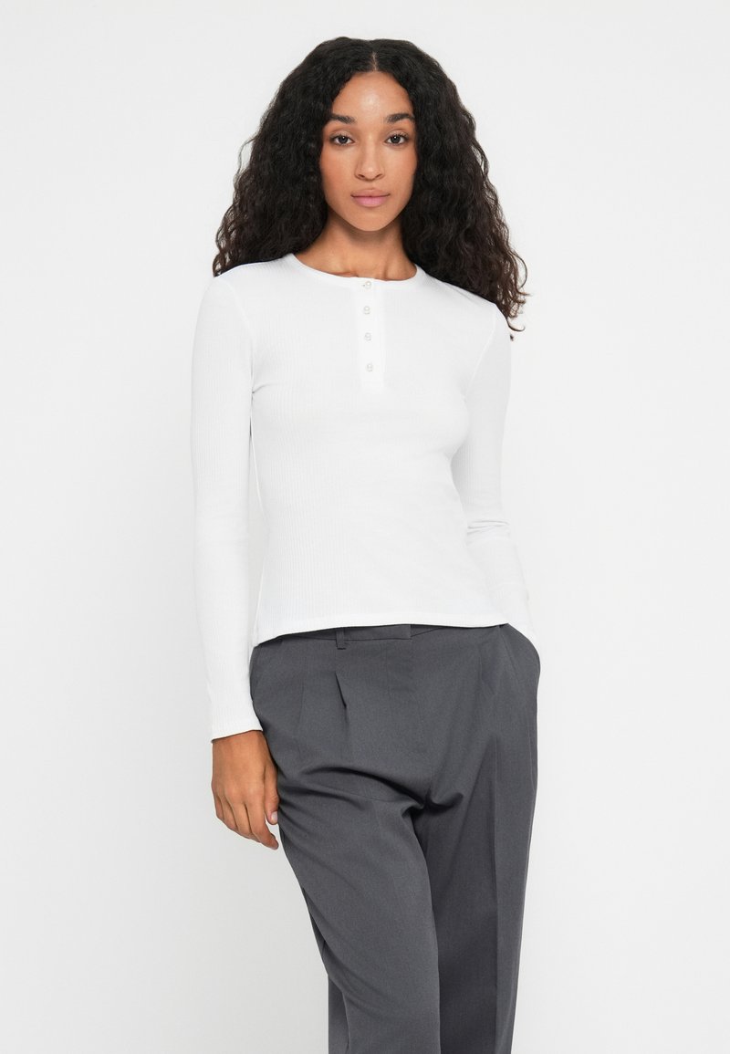 Femme aux longs cheveux bouclés portant un t-shirt ajusté blanc à manches longues avec des boutons et un pantalon plissé gris, se tenant contre un fond blanc.