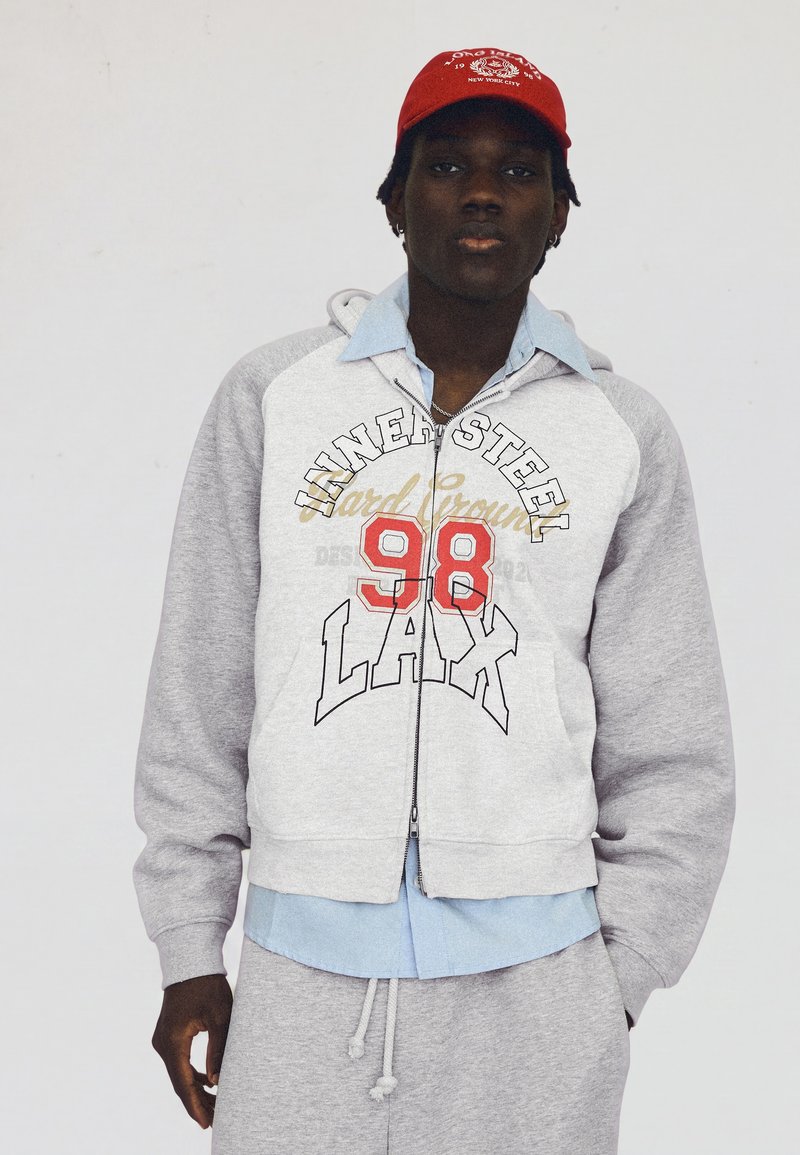 Jeune homme portant un sweat à capuche gris zippé avec le texte "98 LAX", une chemise bleu clair, un pantalon de survêtement gris assorti et une casquette rouge de baseball tournée vers l'avant.
