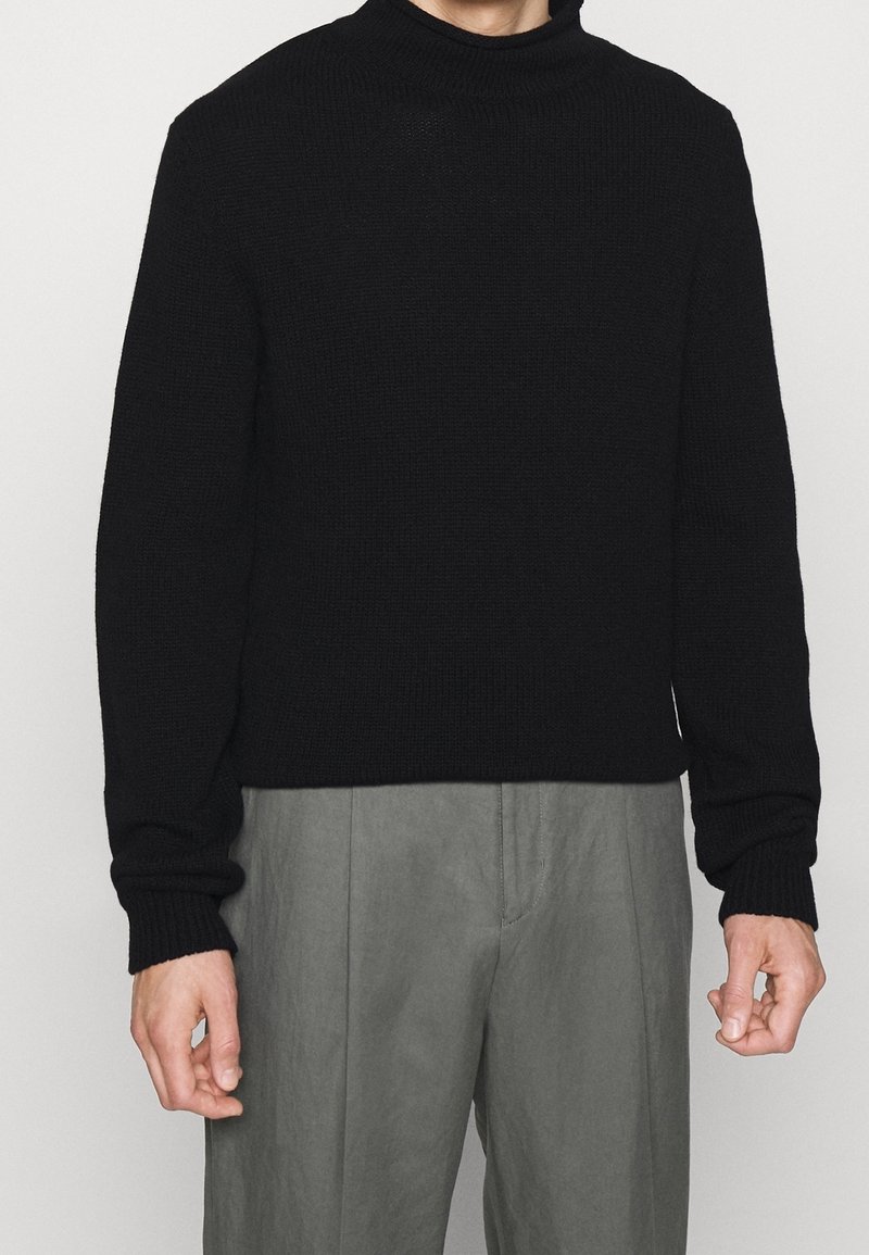 Personne portant un pull noir à col roulé à manches longues et un pantalon gris ample sur un fond uni.