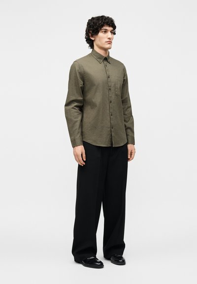 Homme debout portant une chemise vert olive boutonnée, un pantalon noir ample et des chaussures en cuir noires sur un fond clair uni.