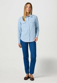 Camisa de manga larga a cuadros azules con dos bolsillos frontales, diseño abotonado, dobladillo redondeado, combinada con vaqueros ajustados azules y zapatos oscuros sin cordones.