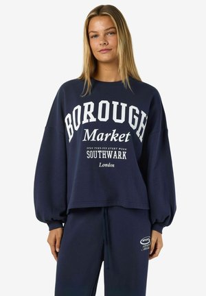 Jeune femme portant un sweat-shirt bleu marine avec le texte "Borough Market Southwark London" et un pantalon de survêtements assorti bleu marine.