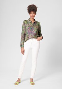Groene paisley-blouse met een kraag, lange mouwen en knoopmanchetten, gecombineerd met witte slim-fit broek en limoen groene sandalen.