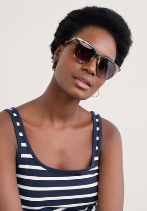Sunglasses with a cat-eye shape, tortoiseshell pattern, dark lenses, and metallic accents. Model wears a navy and white striped tank top.
