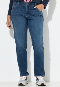 Jeans de mezclilla en azul profundo con una textura suave, diseño de pierna recta y bolsillos delanteros. Llevados con zapatillas grises y azules que presentan acentos en rosa.