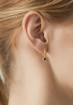 Nærbillede af øre med kort blondt hår iført en lille guldring med en sort charme formet som en firkløver.