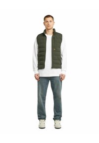 Olivgrön pufferväst med ståkrage, zipförslutning och horisontellt kviltat mönster, bärs över en vit långärmad skjorta och blå jeans.