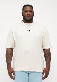 Cream short-sleeve t-shirt in soft fabric, features a butterfly design and "METAMORPHOSIS" text in black on the chest. Relaxed fit.