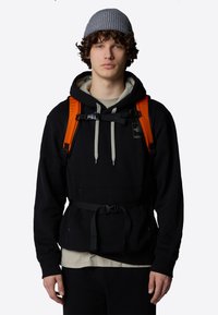 Czarna bluza z kapturem z kieszenią z przodu, w połączeniu z szarym czapką beanie, pomarańczowymi paskami i czarnym paskiem w talii. Warstwowy wygląd obejmuje kremową koszulę pod spodem.