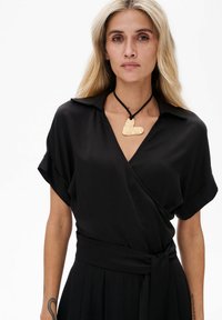 Schwarzes Wickelkleid mit kurzen Ärmeln und Kragen. Mit einer gebundenen Taille und einem goldenen Herzanhänger an einer schwarzen Schnur-Halskette. Glattes Gewebe.