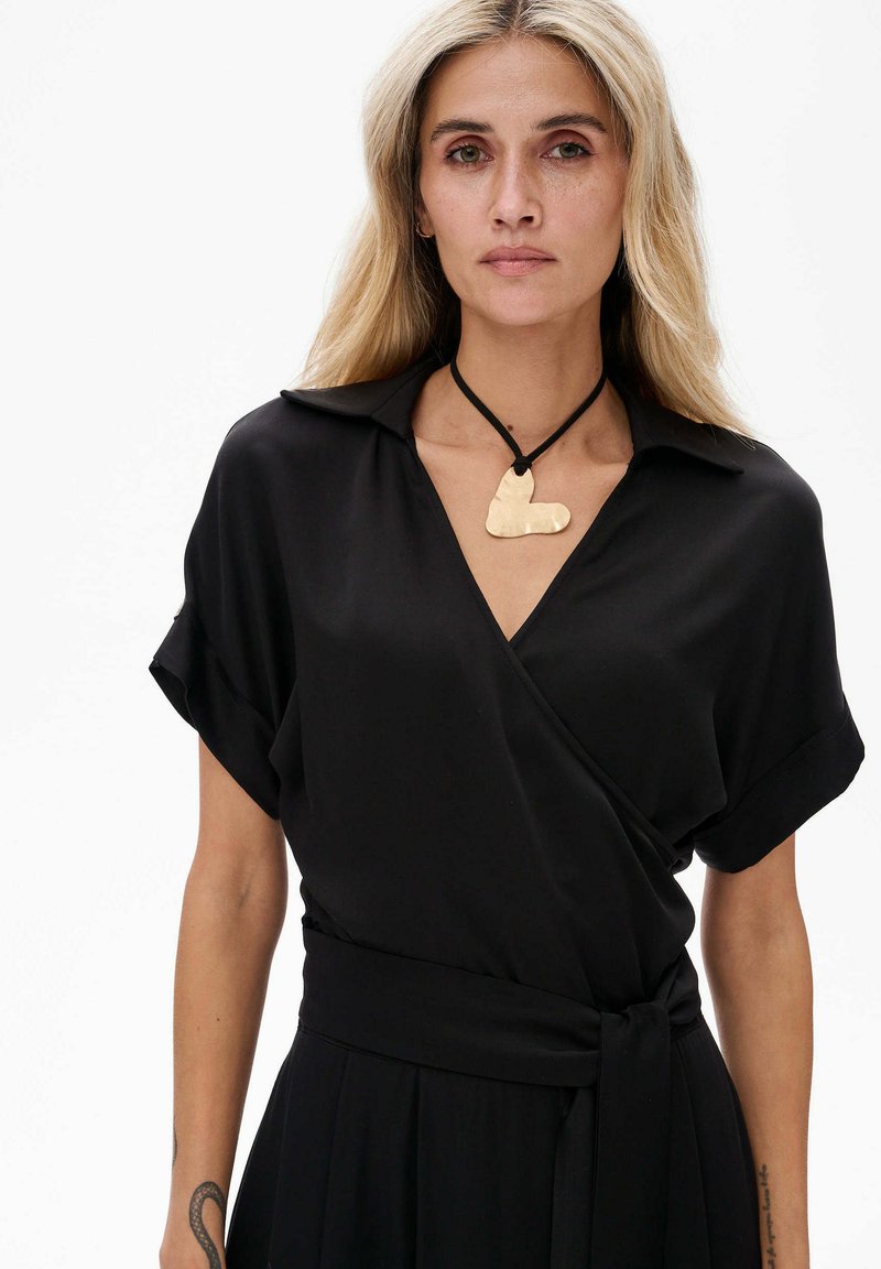 Schwarzes Wickelkleid mit kurzen Ärmeln und Kragen. Mit einer gebundenen Taille und einem goldenen Herzanhänger an einer schwarzen Schnur-Halskette. Glattes Gewebe.