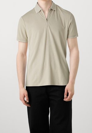 Beige kortærmet polo skjorte med lynlås i halsen og kontrasterende krave. Glat tekstur og en afslappet pasform, kombineret med sorte bukser.