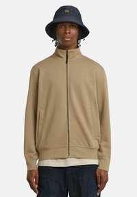 Felpa beige con zip, colletto alto e tasche laterali; indossata sopra una camicia leggera, abbinata a un cappello a secchiello blu con un patch con logo.