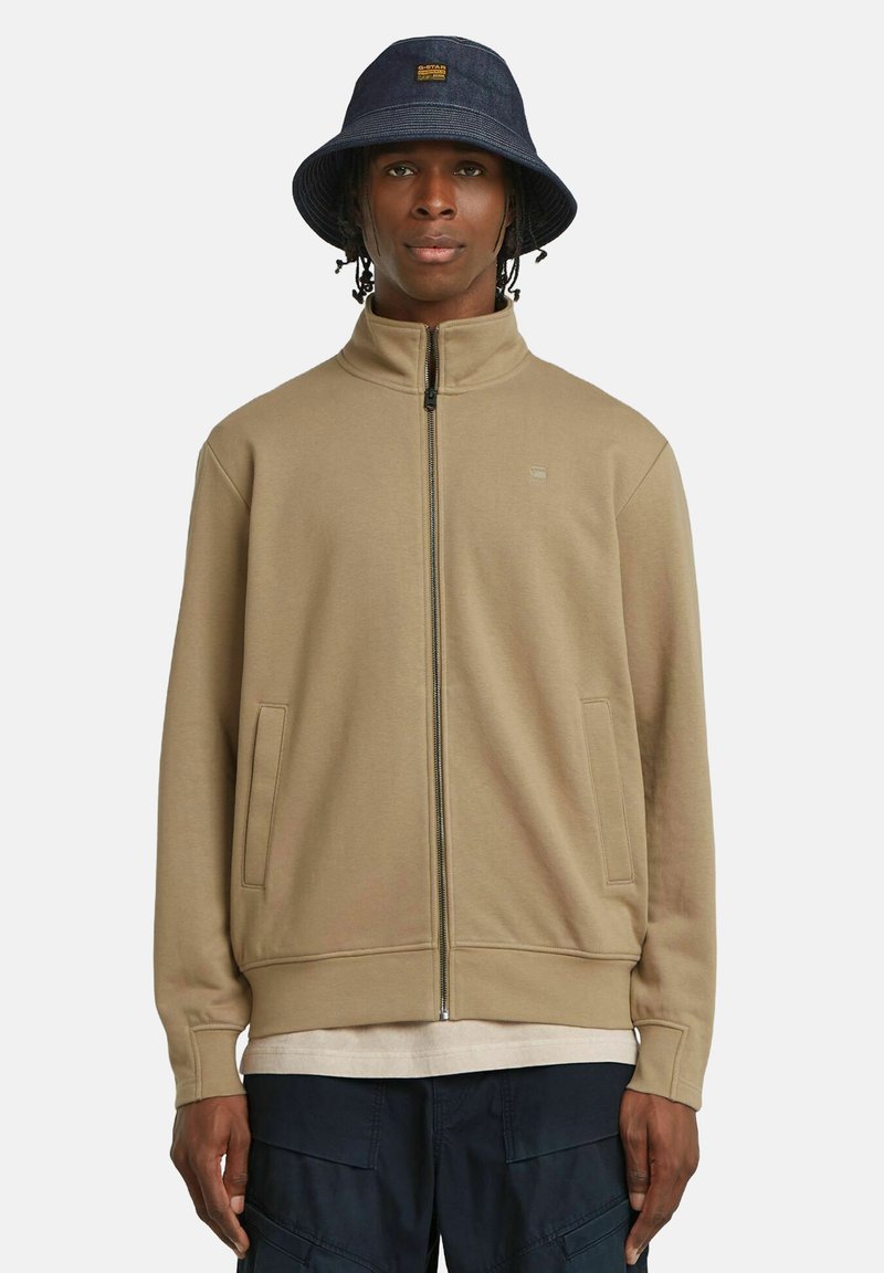 Felpa beige con zip, colletto alto e tasche laterali; indossata sopra una camicia leggera, abbinata a un cappello a secchiello blu con un patch con logo.