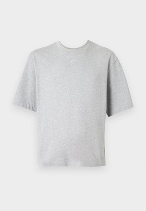 Man staat, draagt een oversized lichtgrijze T-shirt met korte mouwen, zwarte cargobroek, zwarte schoenen en grote futuristische zonnebril, op een grijze vloer.