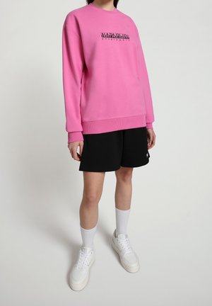 Sudadera rosa con el logo negro "Napapijri Geographic", puños acanalados, llevada con pantalones cortos negros y zapatillas blancas, estilizada con calcetines blancos.