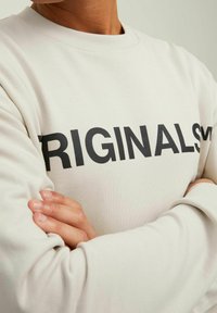 Light beige sweatshirt with black "ORIGINAL" text. Features a crew neck, long sleeves, and ribbed cuffs. Soft cotton material with minimal texture.