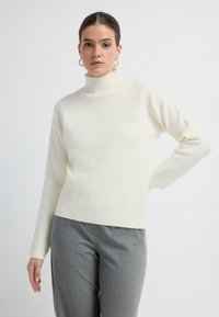 Biały sweter z długim rękawem i golfem, wykonany z miękkiego materiału. Charakteryzuje się dopasowanym krojem i prostym dołem. W zestawie z szarymi spodniami w paski.