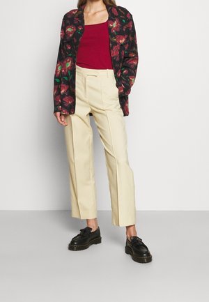 Schwarz mit Blumenmuster bedruckte Jacke, rotes geripptes Tanktop, cremefarbene hochtaillierte Hose und schwarze Schnürschuhe mit dicker Sohle.