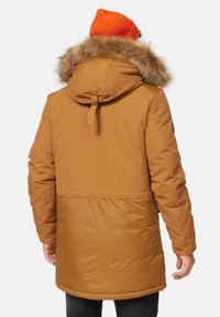 Parka marron avec une capuche doublée de fourrure et un cordon de serrage, présentant une texture lisse et une finition légèrement brillante. Patch noir sur le bras visible.