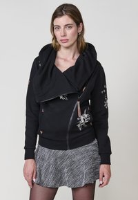 Sudadera negra texturizada con un gran cuello doblado, cremallera lateral y bordado floral. Presenta acentos marrones y se combina con una falda gris.