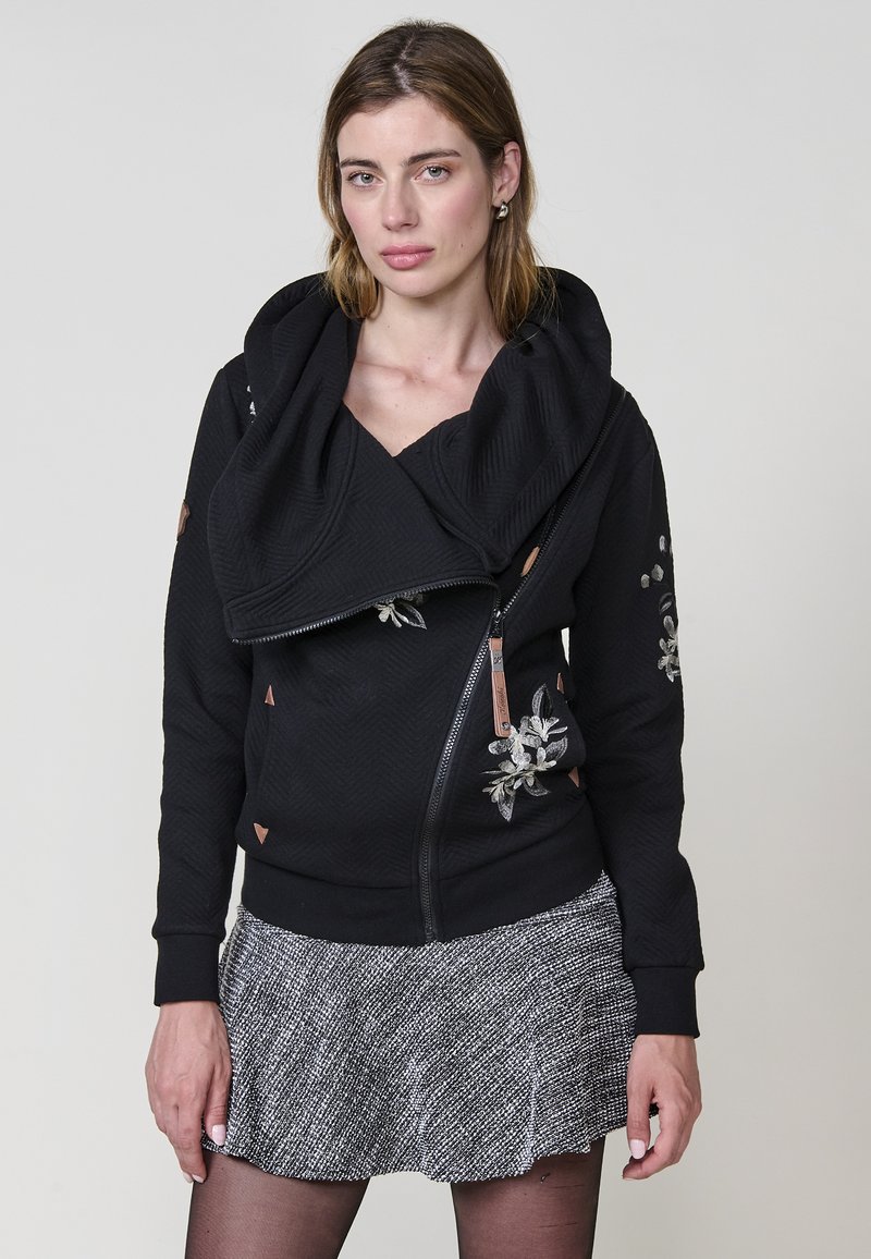 Sudadera negra texturizada con un gran cuello doblado, cremallera lateral y bordado floral. Presenta acentos marrones y se combina con una falda gris.
