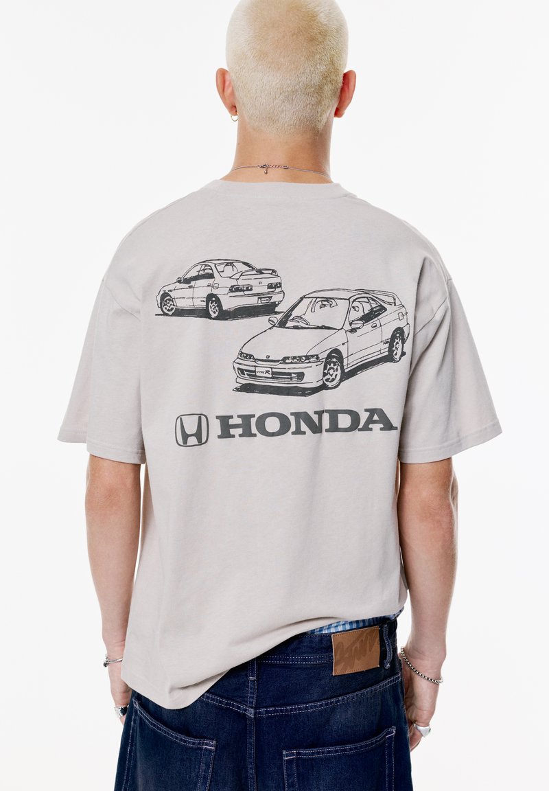 Lys beige bomulds t-shirt med sorte skitser af to biler og et "Honda" logo på ryggen. Oversized pasform, korte ærmer.