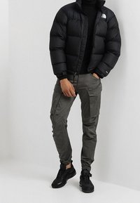 Zwarte pufferjack met een gewatteerd patroon, donkergrijze cargobroek met zijzakken, en zwarte sneakers met een gladde afwerking.