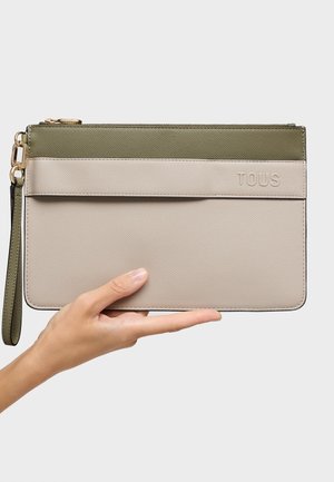 Mano che tiene una pochette da polso rettangolare bicolore con la parte superiore verde oliva e la parte inferiore beige, con il nome del marchio TOUS in rilievo.