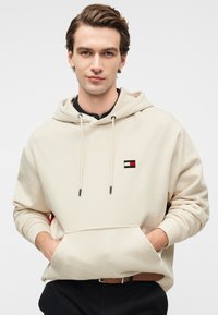 Μπεζ φούτερ με κουκούλα, μπροστινή τσέπη, μαύρα κορδόνια και μικρό κεντητό λογότυπο σε κόκκινο, άσπρο και μαύρο στο στήθος. Ομαλή υφή.