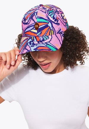 Femme aux cheveux bouclés portant un haut blanc et une casquette de baseball rose avec des motifs colorés de papillons, ajustant la visière de la casquette.