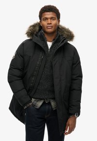 Chaqueta de plumas negra con capucha forrada de pelo, cierre de cremallera, bolsillos laterales, llevada sobre un suéter oscuro y una camisa de cuadros. Jeans y reloj visibles.