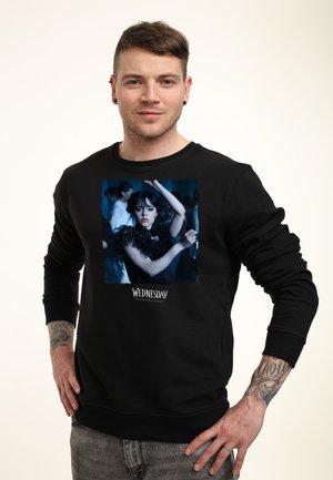 Mand iført en sort sweatshirt med et billede af en kvinde i en mørk kjole med en hævet arm og teksten "Wednesday" nedenfor billedet.