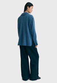Camisa azul de manga larga con cuello, que presenta un pliegue en la parte posterior, combinada con pantalones oscuros de pierna ancha. Tejido suave con un ajuste relajado.