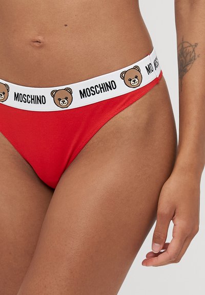 Sarkans kokvilnas stringi ar baltu elastīgu jostasvietu, uz kuras ir drukāts lācis un teksts "Moschino", izceļot gludu tekstūru un minimālistisku dizainu.