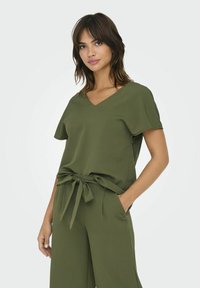 Top de color verde oliva, de manga corta con escote en V y cintura anudada. La tela parece suave y texturizada, combinada con pantalones sueltos a juego.