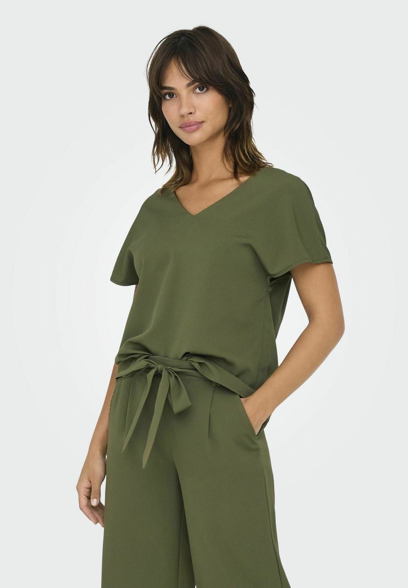 Top de color verde oliva, de manga corta con escote en V y cintura anudada. La tela parece suave y texturizada, combinada con pantalones sueltos a juego.